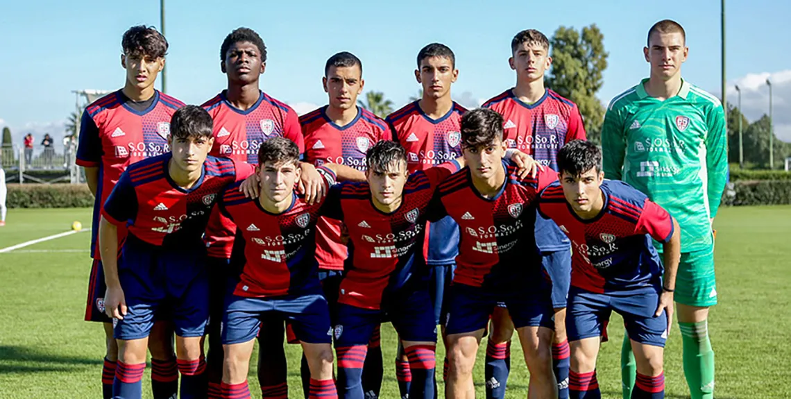 Foto di gruppo della squadra under 17 del Cagliari calcio. I ragazzi sono disposti su due file, da 6 e 5 persone. I 5 ragazzi della fila davanti sono chinati, i 5 ragazzi dietro sono in piedi. Indossano la divisa rossoblu, tranne il portiere in alto a destra che ha una divisa verde.