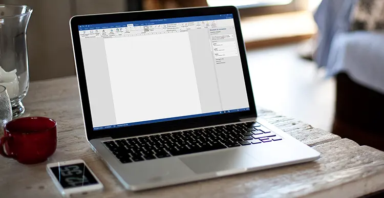 Laptop con schermata Microsoft Word, poggiato su un tavolo di legno. Alla sinistra del laptop sono poggiati una tazza rossa e uno smartphone.