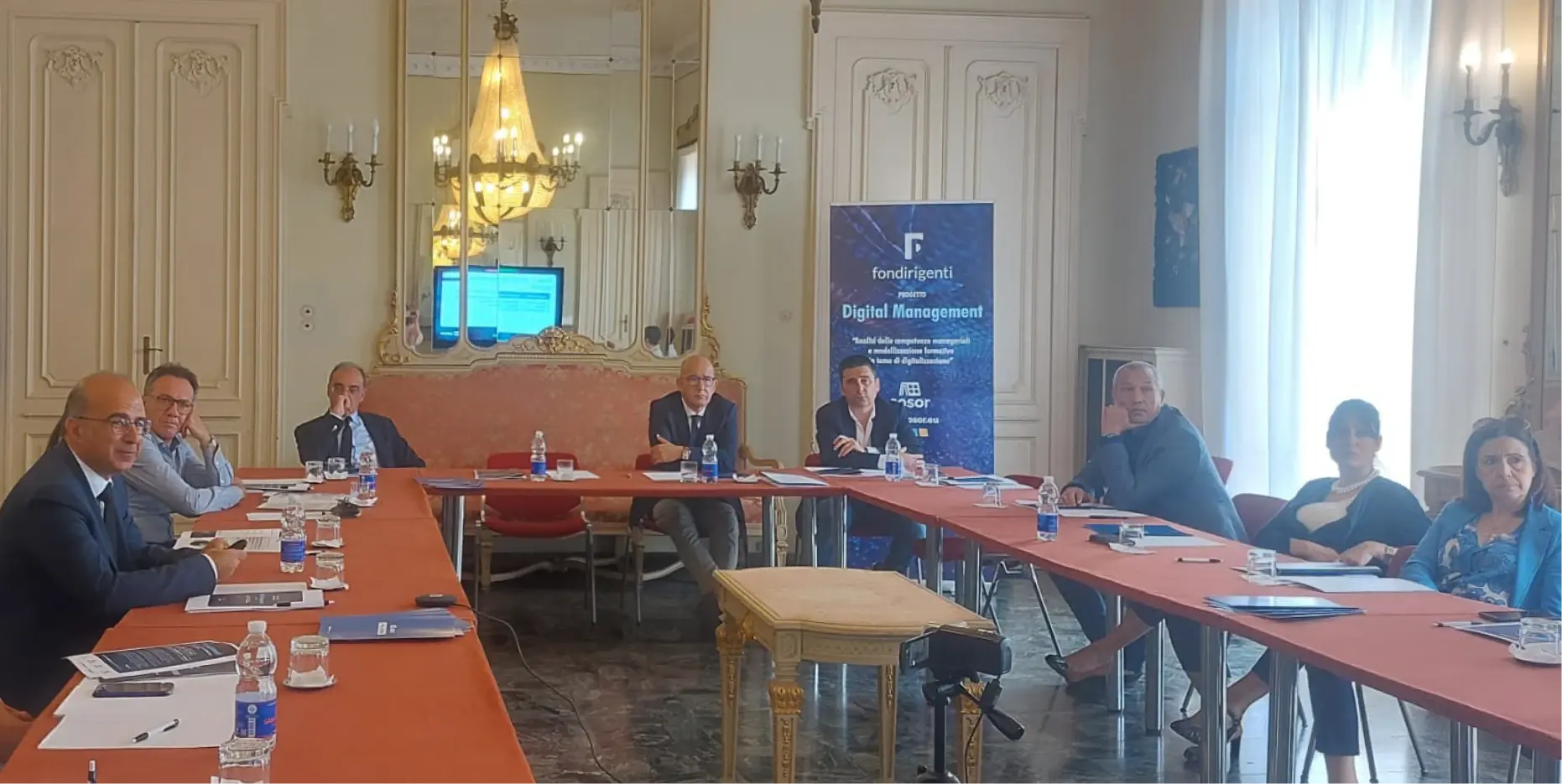 Otto persone, sei uomini e due donne, sedute intorno a tavoli disposti a formare un rettangolo, in una sala in stile Rococò con specchi e lampadari di cristallo