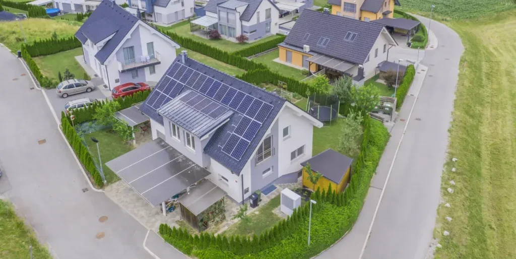 Veduta rappresentativa del progetto CER Sardegna. Un gruppo di case bianche viste dall'alto, circondate da prati. Il tetto della casa al centro è coperto da pannelli fotovoltaici.