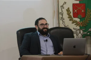 R.Raccis, sorridente. Ha corti capelli scuri e ricci, barba media, occhiali scuri e giacca scura su camicia azzurra