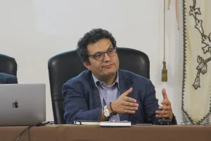 Avv.to Michele Loche. Ha corti capelli ricci scuri e occhiali, indossa giacca blu e camicia azzurra