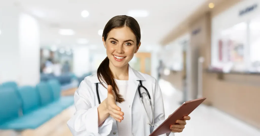 Nuove disponibilità corso OSS a Sorso - Giovane donna sorridente che porge la mano. Ha i capelli castani lisci raccolti, indossa camice bianco e stetoscopio e regge una cartella. Alle sue spalle, un corridoio di una struttura sanitaria.