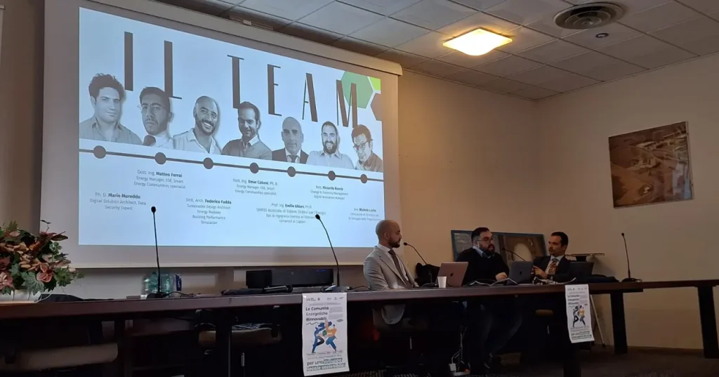 Villacidro, un momento del 6 seminario CER Sardegna. Tre relatori sono seduti a un lungo tavolo, alle loro spalle è proiettata una foto del gruppo di lavoro, composto da 7 uomini.