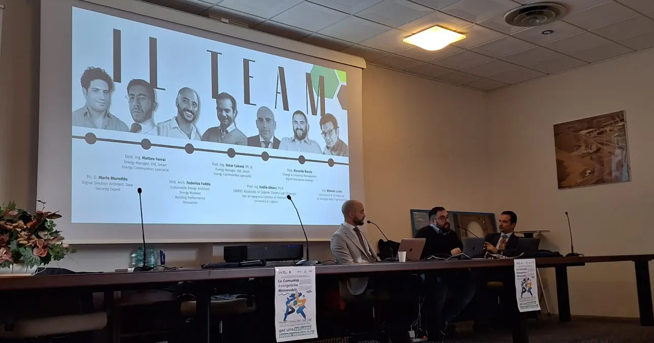 Villacidro, un momento del 6 seminario CER Sardegna. Tre relatori sono seduti a un lungo tavolo, alle loro spalle è proiettata una foto del gruppo di lavoro, composto da 7 uomini.