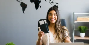 Una ragazza mora con i capelli lunghi, seduta, sorride mostrando due biglietti. Alle sue spalle una parete con parte del planisfero che mostra America Latina, Africa e Australia - Corso gratuito Destination Manager - Prato, Toscana