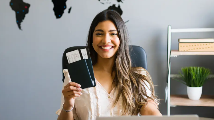 Una ragazza mora con i capelli lunghi, seduta, sorride mostrando due biglietti. Alle sue spalle una parete con parte del planisfero che mostra America Latina, Africa e Australia - Corso gratuito Destination Manager - Prato, Toscana