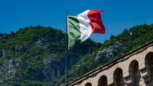Bandiera italiana su pennone sventola in aria, sullo sfondo una collina verde e un edificio antico