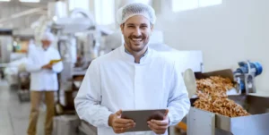 Giovane con camice bianco e retina nei capelli tiene un tablet dentro una fabbrica di alimenti