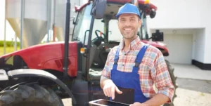 Uomo in salopette da lavoro e casco antinfortunistico tiene in mano un tablet e sorride davanti a un trattore