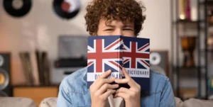 Giovane coi capelli ricci regge un dizionario di inglese che gli copre il viso sino a metà naso