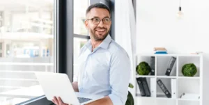 Uomo in camicia e occhiali regge un computer portatile in un ufficio e sorride