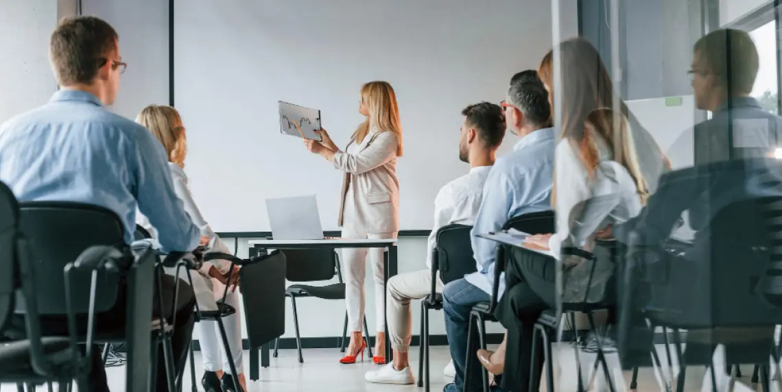 Gruppo di donne e uomini in un'aula segue una lezione tenuta da una donna bionda che mostra un grafico