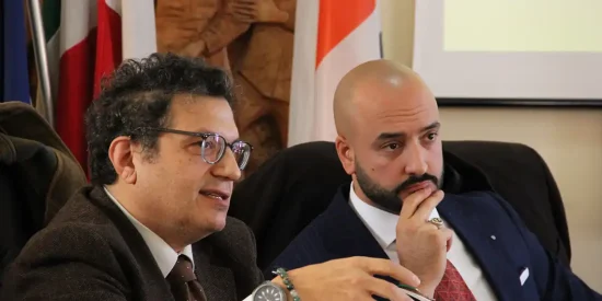 Michele Loche e Federico Fadda in giacca scura, cravatta rossa e camicia bianca. Sono seduti, Loche porta gli occhiali e i capelli corti, Fadda ha la testa rasata e la barba